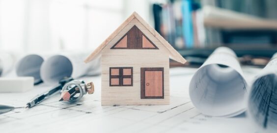 A small, wooden model house is sitting on top of a paper, alongside rolled up blueprints and drawing tools.