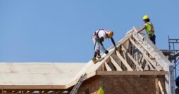 Two men in brightly colored safety gear work on a residential roof with exposed wood beams and visible roof decking.