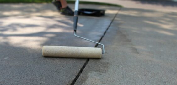 A paint roller in focus as a person in the background rolls sealant onto a concrete driveway, darkening the surface.