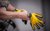 A contractor places work gloves on their hands. The work gloves are yellow with black support features on the fingers.