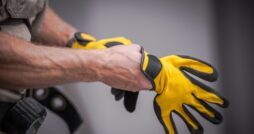 A contractor places work gloves on their hands. The work gloves are yellow with black support features on the fingers.