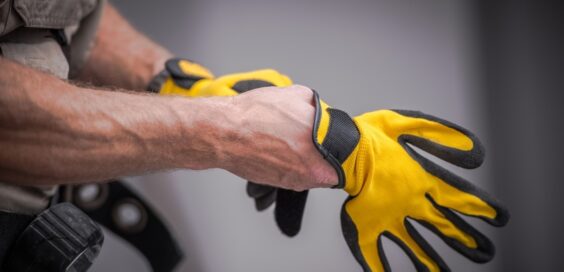 A contractor places work gloves on their hands. The work gloves are yellow with black support features on the fingers.