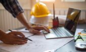 A close-up of a person wearing a flannel shirt, holding a pencil, and looking at plans. They are also using a laptop.