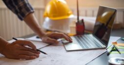 A close-up of a person wearing a flannel shirt, holding a pencil, and looking at plans. They are also using a laptop.