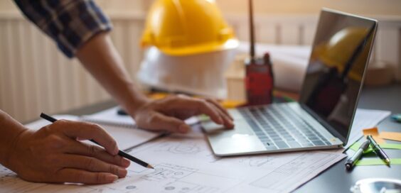 A close-up of a person wearing a flannel shirt, holding a pencil, and looking at plans. They are also using a laptop.