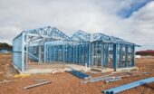 A house that is in the process of being built on an empty lot appears to have a frame made out of a blue colored metal.