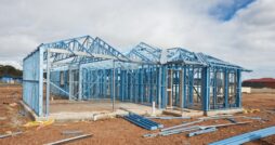 A house that is in the process of being built on an empty lot appears to have a frame made out of a blue colored metal.