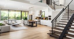 A home with an open-concept layout includes a staircase with black handrails, white cabinets in the kitchen, and a gray sofa.