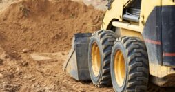 A yellow skid steer using a bucket attachment to push dirt into a pile.
