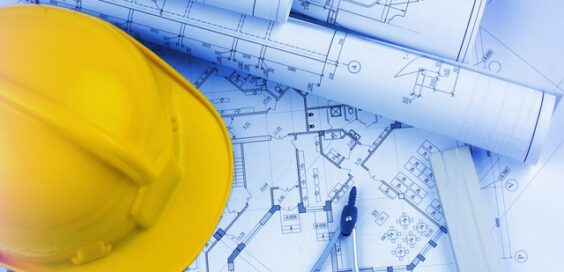 A close-up of construction blueprints lying on the table with a yellow hard hat with a compass sitting on top.