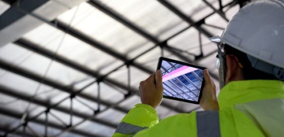 A man in PPE points a tablet up toward the ceiling of an industrial facility. The tablet highlights a ceiling beam in pink.