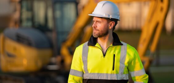 A man in a high-visibility top with a white hard hat looks into the distance as he stands in front of a blurred excavator.