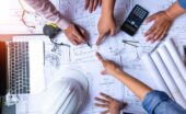 Three people stand around a table reviewing housing plans, pointing at them beside a hard hat, laptop, tape, and calculator.