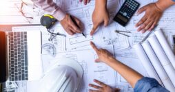 Three people stand around a table reviewing housing plans, pointing at them beside a hard hat, laptop, tape, and calculator.