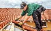 A roofing contractor standing on a roof and placing layers of red-brown terra cotta tiles against a wooden frame.
