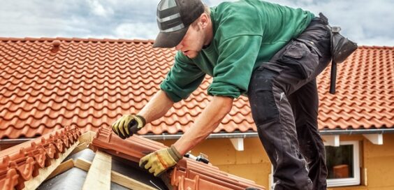 A roofing contractor standing on a roof and placing layers of red-brown terra cotta tiles against a wooden frame.