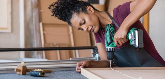 A close-up of a woman with brown curly hair assembling a frame with a drill in her personal workshop.