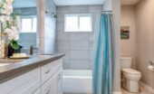 The interior of a warm bathroom space. The prominent colors are white and gray and there is a blue shower curtain.