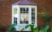 A bay window with white frames attached to a brick house. Trees are reflected in the glass, and plants are growing under it.