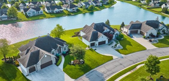 Aerial view of a suburban neighborhood with large homes, green lawns, trees, and a winding lake under bright daylight.