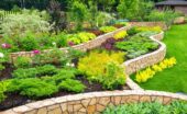 Tiered stone retaining walls create colorful garden beds on a sloped lawn with vibrant plants and flowers at each level.