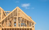 The wooden frame of a large house under construction, featuring an A-frame roof against a bright blue sky.