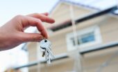 A close-up of a hand holding a ring of keys with the thumb and index finger with a home blurred in the background.
