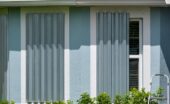Gray, metal hurricane shutters are covering the windows on a light-blue, stucco home. Small bushes appear under the windows.