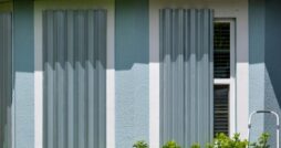 Gray, metal hurricane shutters are covering the windows on a light-blue, stucco home. Small bushes appear under the windows.