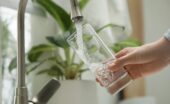 A close-up view shows a person holding a tall, clear glass under a stainless steel faucet and filling it with water.