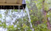 A roof gutter overflows with water during a storm, with splashing droplets and green trees in the background.