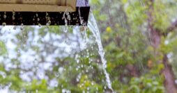 A roof gutter overflows with water during a storm, with splashing droplets and green trees in the background.