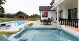A backyard patio with a hot tub beside a modern home, pool, red chairs, and a covered gazebo on an overcast day.