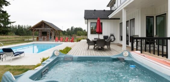A backyard patio with a hot tub beside a modern home, pool, red chairs, and a covered gazebo on an overcast day.