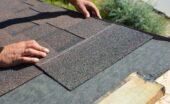 A close-up of a contractor's hands laying down an asphalt shingle marked with a white line onto a roof's underlayment.