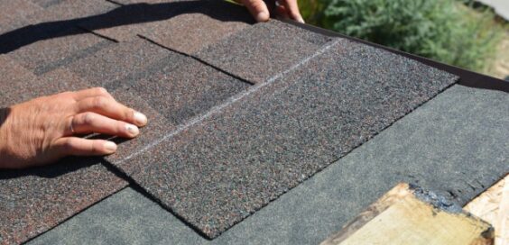 A close-up of a contractor's hands laying down an asphalt shingle marked with a white line onto a roof's underlayment.
