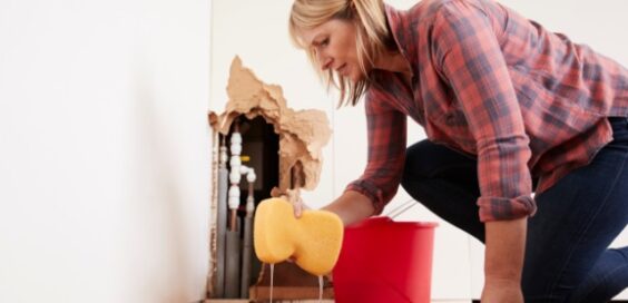 A woman using a sponge to mop up a large puddle that has come from a hole in the wall, exposing the plumbing within.