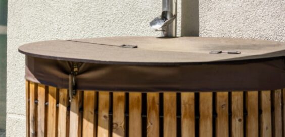 A close-up of a rainwater harvesting barrel stored outside a home with a metal downspout leading down from the roof.