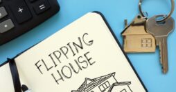 A close-up of a notepad open on a blue surface with the words "Flipping House" written next to a set of keys.