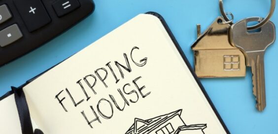 A close-up of a notepad open on a blue surface with the words "Flipping House" written next to a set of keys.