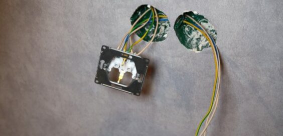 Partly installed electrical socket dangles from exposed wires beside two circular openings in a textured gray wall.