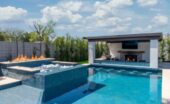 A large backyard with a clear pool, fire features, outdoor grill, and wall-mounted screen. The sky above is cloudy.
