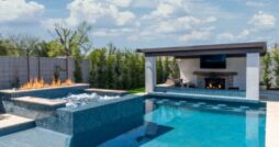 A large backyard with a clear pool, fire features, outdoor grill, and wall-mounted screen. The sky above is cloudy.