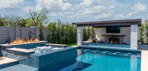 A large backyard with a clear pool, fire features, outdoor grill, and wall-mounted screen. The sky above is cloudy.