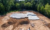 A concrete foundation slab is set on a cleared construction site surrounded by dirt and trees before building begins.