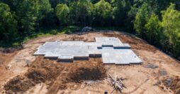 A concrete foundation slab is set on a cleared construction site surrounded by dirt and trees before building begins.