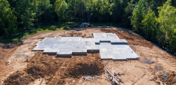 A concrete foundation slab is set on a cleared construction site surrounded by dirt and trees before building begins.