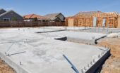 A concrete home foundation sits at a construction site beside wooden framing and nearby houses under a blue sky.
