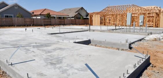 A concrete home foundation sits at a construction site beside wooden framing and nearby houses under a blue sky.