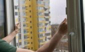 A person holding a mesh screen frame in an open window, with distant buildings visible through the net.